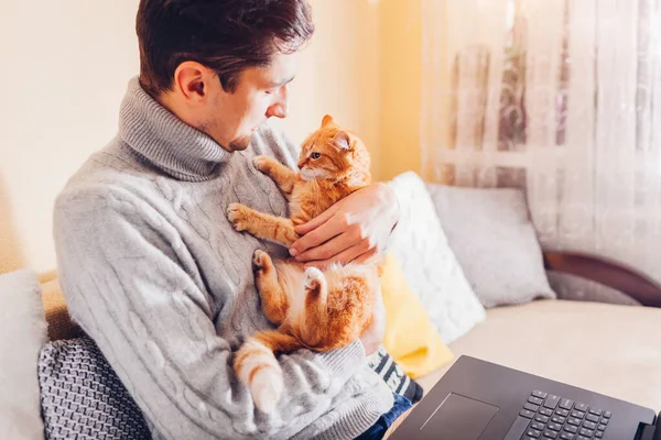 Man holding cat working in internet from home using laptop. Young freelancer playing with pet sitting on couch. Remote job
