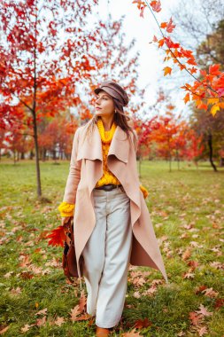 Güz parkında kırmızı ağaçların arasında şık, sıcak sarı, bej kıyafetler giyen güzel bir kadının vücut portresi. Kadın sonbahar modası. Modern aksesuarlar