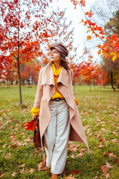 Güz parkında kırmızı ağaçların arasında şık, sıcak sarı, bej kıyafetler giyen güzel bir kadının vücut portresi. Kadın sonbahar modası. Modern aksesuarlar
