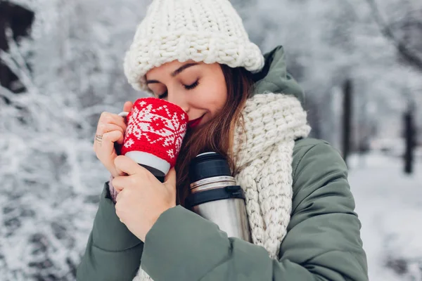 Woman drinking hot tea holding vacuum flask in snowy winter park enjoying landscape under falling snow. Cup dressed in red knitted Christmas case