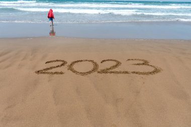 Yıl 2023, kumsalda, kırmızı ceketli bir çocukla ve arka planda denizle birlikte yazılmış..