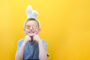 Gözlüklü bir çocuk bir Paskalya tavşanının kulaklarını takmış ve lolipop veya şeker yiyor.