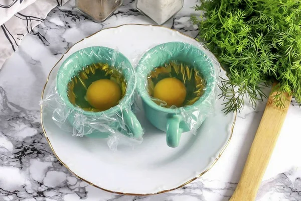 Cut a piece of cling film into two wide squares. Brush them with vegetable oil and place in cups with the oiled side up. Crack an egg into the bowl.