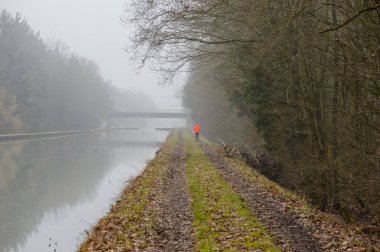 Alsace France 'da koşucu. Sabah sisinde Canal de la Marne au Rhin. Kırmızı anorak 'lı bir koşucu.