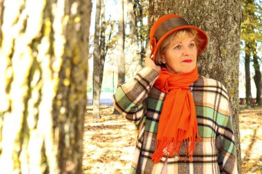 Eski parkta sonbahar yürüyüşleri: turuncu şapkalı ve ekose ceketli yaşlı bir kadın şapkasını düzelterek eski bir ağacın yanında duruyor.