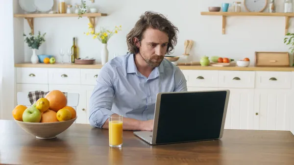 Vonzó férfi, aki laptoppal dolgozik otthonról. Egy gondolkodó ember ül az asztalnál, iszik egy üdítőt a pohárból, miközben távolról dolgozik otthonról. A távmunka problémájára való reflektálás — Stock Fotó