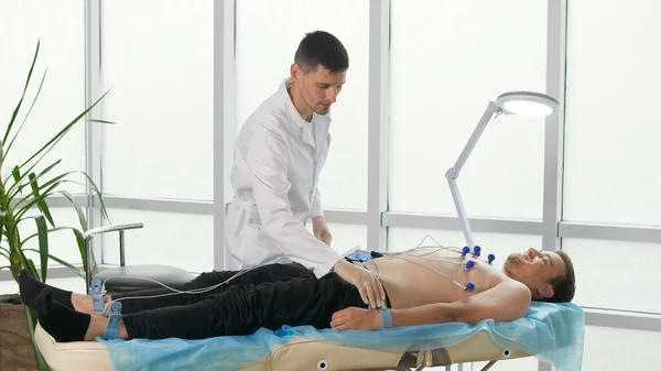 心臓病の診断のための心電図手順.心臓専門医が若い男の胸の上に電極を置き、診療所で心電図を取るためにソファに寝そべって. — ストック写真