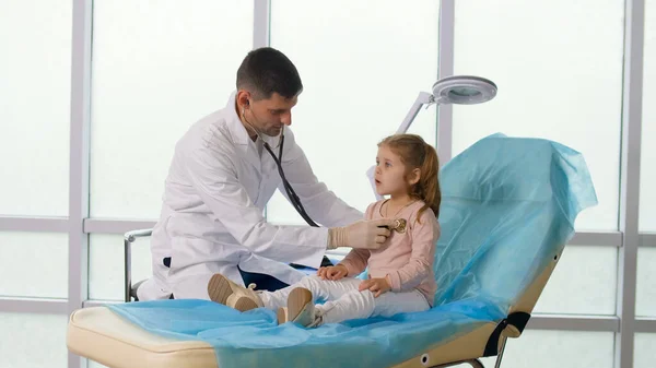 Пациент посещает врачебный кабинет. Доктор слушает легкие ребёнка со стетоскопом, говорит с маленькой девочкой заботливо. Удобное пребывание ребенка в клинике. — стоковое фото