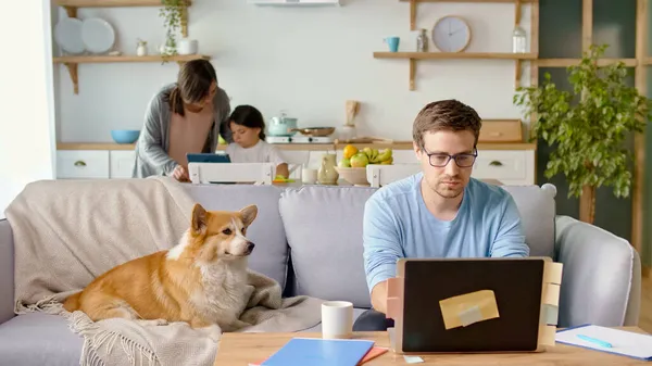 Elfoglalt apa Távolról dolgozik Laptop ül a konyhában. Egy aranyos kiskutya fekszik a kanapén a közelben. A háttérben a felesége segít a lányának online tanulni a tabletjén. Maradj a házban. — Stock Fotó