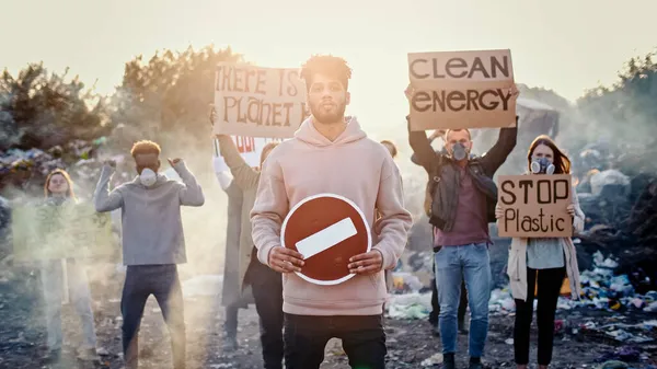 Portrét atraktivního mladého aktivisty, který drží stopku. V pozadí lidé protestují proti znečišťování odpadků. Koncepce změny klimatu. Royalty Free Stock Obrázky