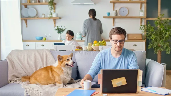 Elfoglalt apa Távolról dolgozik Laptop ül a konyhában. Egy aranyos kiskutya fekszik a kanapén a közelben. A háttérben a felesége segít a lányának online tanulni a tabletjén. Maradj a házban. — Stock Fotó