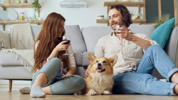 Happy Young Married Couple Sitting on the Floor Near a Couch and Clinking Glasses With Wine