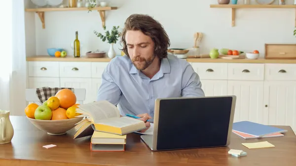 A mai generáció távolról tanulhat otthonról. Vonzó Man használ könyveket és laptopot tanulásra otthon. Egy férfi jegyzetel a füzetben. — Stock Fotó