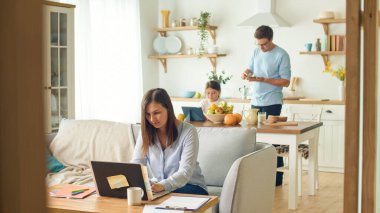 Annem dizüstü bilgisayarla uzaktan kumandayla koltukta oturmakla meşgul. Karantina salgını sırasında evden çalışmak. Arka planda, kızı bir tablet kullanıyor, kocası da mutfakta çok meşgul.