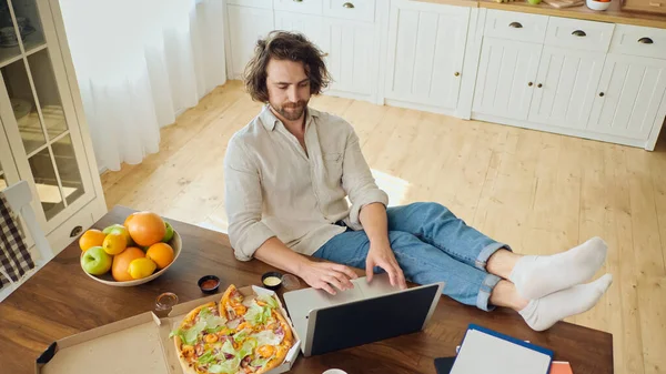 Vonzó fáradt férfi dolgozik egy laptop otthonról. Férfi ül az asztalnál Egyél egy pizzát közben távolról dolgozik otthonról. — Stock Fotó