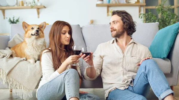 Happy Young Married Couple Sitting on the Floor Near a Couch and Clinking Glasses With Wine