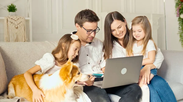 Een gelukkig gezin maakt gebruik van een laptop voor online winkelen, Zittend op de bank thuis. Kopen via het internet. Een familly zittend met een schattige hond op de bank thuis. Aankoopbevestiging via internet Stockfoto