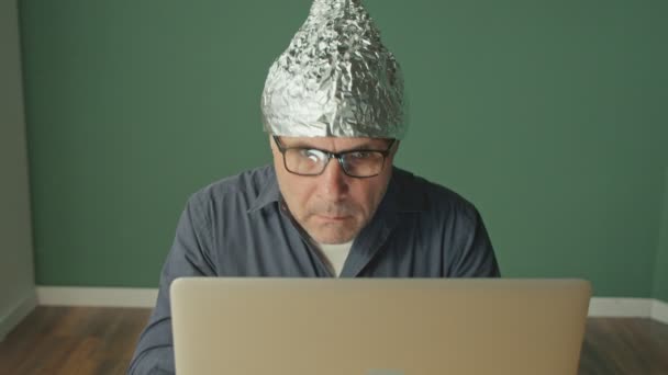 Un hombre asustado en una tapa de papel de aluminio sentado en una computadora portátil, mirando ansiosamente a su alrededor. — Vídeo de stock