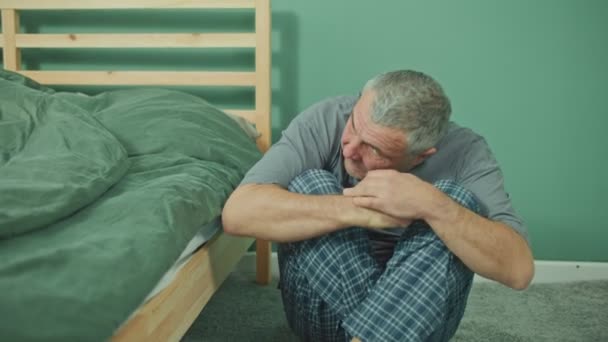 En rädd man sitter på sovrumsgolvet bredvid sängen, mental hälsa. — Stockvideo