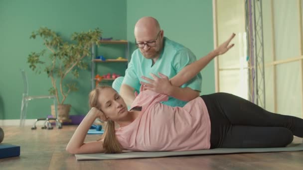Fizjoterapeuta pomaga dziewczynie zrobić ćwiczenia łagodzić ból w klinice odwykowej. — Wideo stockowe