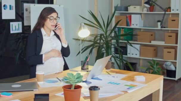 Una hermosa mujer embarazada hablando en un teléfono celular sentada en una mesa — Vídeo de stock