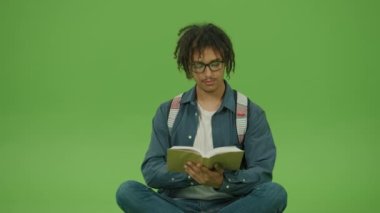 Gözlüklü ve Denim Shirt giyen genç Meksikalı adam kitap arıyor, sayfaları çeviriyor ve okumanın tadını çıkarıyor. Yeşil Ekran Arkaplanına Karşı Kitap Oturan Yakışıklı Motivasyonlu Adam Portresi