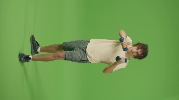 縦の映像 スポーツはベージュのシャツで若いメキシコ人を動機づけ緑の画面の背景に対してダンベルとボクシングによって演習を行う 強さとホームワークアウト 国内物理的ジャーク — ストック動画