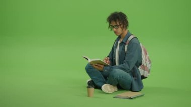Gözlüklü ve Denim Shirt 'lü Genç Çok Kültürlü Adam Kitap ve Gülümseyen Sayfalar Arıyor. Yeşil Ekran Arkaplanında Kitaplarla, Notlarla ve Kahveyle oturan Yakışıklı Adam 'ın Portresi