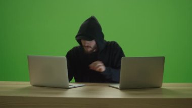 Tanınmayan Adam Korsanının Başlıklı ve Kapşonlu Tişörtü Giymesi Ahşap Masadaki 2 Laptop Bilgisayar Klavyesine Yazması. Tehlikeli Kapüşonlu Bilgisayar Korsanının Yeşil Ekran 'da Siber Sistemleri Kırması, Krom Anahtar Sistemi