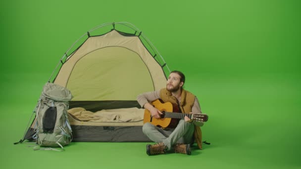 夢のような若いひげを生やした男バックパックとギターを演奏してオープンテントの近くに座って 星を見て 緑の画面で満足笑みを浮かべて クロマキー 旅行とアドベンチャーのコンセプト — ストック動画