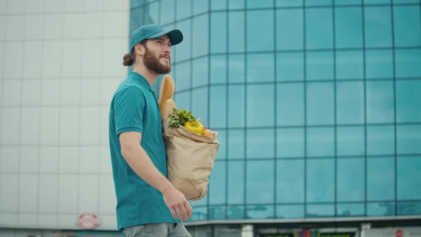 制服の配達人は食品と紙袋を保持している背の高いガラス建築ビジネスセンターの間で小包を届けに行く 市内のフード パーツの迅速な宅配 — ストック動画