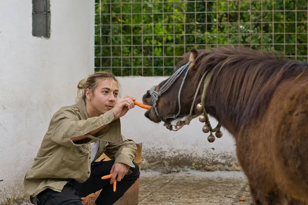 Uzun sarı saçlı genç bir adam midilliyi besliyor.