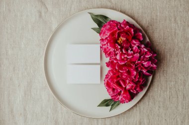 Şakayık çiçekleri ve tomurcuklarla açan dallar. Seramik tabaktaki boş kartvizit modeli. Kadınsı düğün ve kırmızı şakayık çiçekleri. Gri keten masa örtüsü kumaş arka plan. Botanik ve lüks