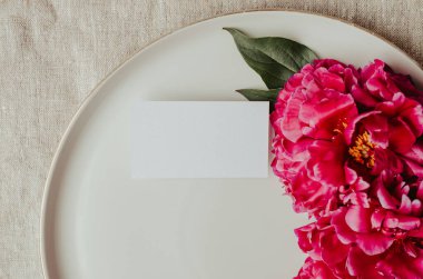 Şakayık çiçekleri ve tomurcuklarla açan dallar. Seramik tabaktaki boş kartvizit modeli. Kadınsı düğün ve kırmızı şakayık çiçekleri. Gri keten masa örtüsü kumaş arka plan. Botanik ve lüks