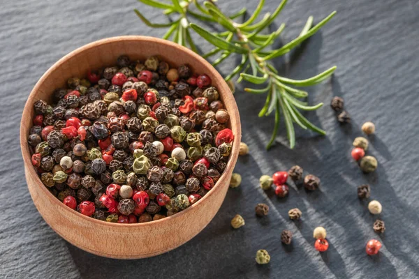 Peppercorns seeds of different colors in wooden plate closeup, black background — Foto Stock