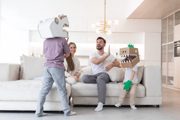 家庭で恐竜を遊んでいる両親と2人の子供の幸せな家族 段ボールの手作りの衣装マスクを身に着けている子供 — ストック写真