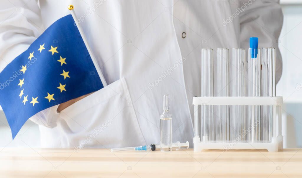 Doctor stands near test tube, ampoule, syringe and flag of Europe Union, concept of global vaccination.