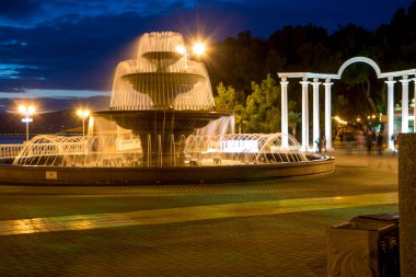 Üç katmanlı fıskiyeler ve günbatımı gökyüzünün arka planında sütunları olan kemerler söylüyor. Akşam Gelendzhik seti. Işıklar kirişlerle dolgu seti üzerinde parlıyor. Siluetler