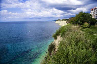 High rocky coast of the Black Sea. High layered rocks. Lighthouse on the seashore. Blue sea summer, white clouds.