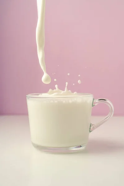 Mleko wlewa się do przezroczystego kubka na fioletowym tle. — Zdjęcie stockowe