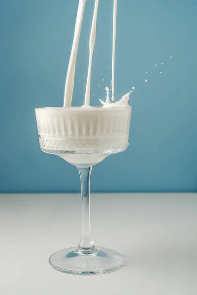 La leche se vierte en un hermoso vaso de cristal sobre un fondo azul y salpica alrededor. — Foto de Stock