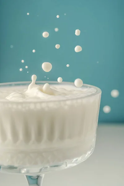 Mleko wlewa się do pięknej kryształowej szklanki na niebieskim tle i rozbryzga się wokół. — Zdjęcie stockowe
