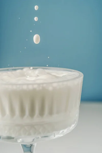 La leche se vierte en un hermoso vaso de cristal sobre un fondo azul y salpica alrededor. — Foto de Stock