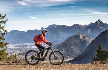 Doğu Allgaeu dağlarında elektrikli dağ bisikletine binen yaşlı güzel bir kadın. Arka planda Zugspitze Dağı, Bavyera Alpleri, Almanya