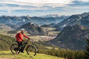 Doğu Allgaeu dağlarında elektrikli dağ bisikletine binen yaşlı güzel bir kadın. Arka planda Zugspitze Dağı, Bavyera Alpleri, Almanya
