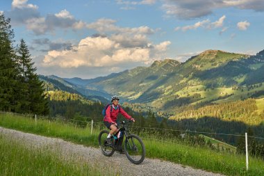 pretty senior woman riding her electric mountain bike on the mountains above Oberstaufen with Nagelfluh mountain chain in background, Allgau Alps, Bavaria Germany clipart
