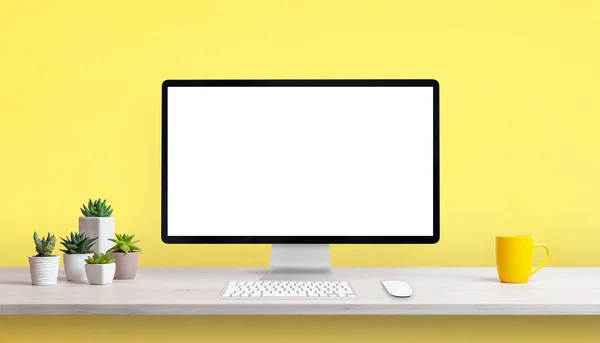Creative work desk with blank computer display, yellow coffee mug and plants. Yellow background with copy space