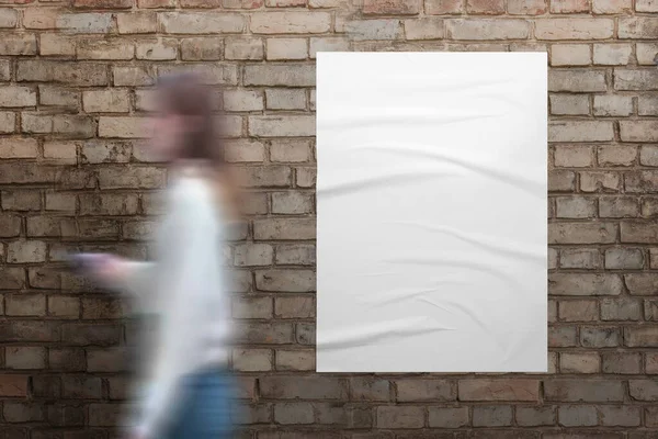 女の子はレンガの壁に白いポスターを通り過ぎる デザインプレゼンテーションのためのしわのテクスチャを持つ空白の白いポスター — ストック写真
