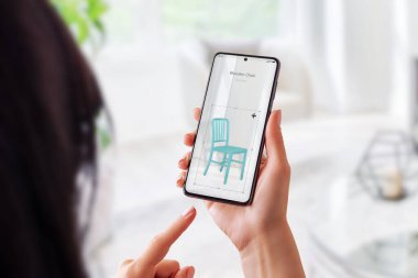 Arttırılmış gerçeklik uygulaması olan bir kadın oturma odasında tahta bir sandalye kuruyor. Mobil konsepte sahip sanal mobilya yerleştirme
