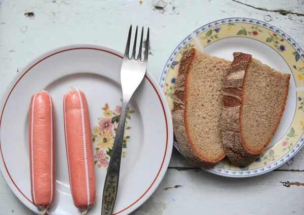 Bir tabakta iki sosis, beyaz ahşap bir masada iki dilim siyah ekmek.